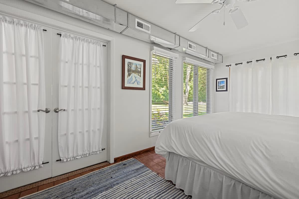 Bedroom with a white bed, terracotta tile flooring, and double French doors leading outside. Windows with blinds offer views of the exterior, and a ceiling fan is visible.