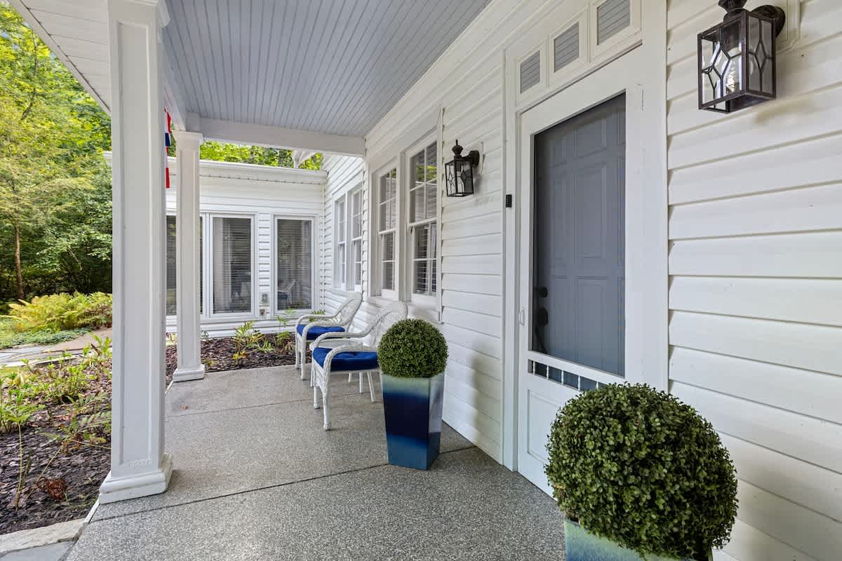 A welcoming front porch features a concrete floor, white siding, and two white chairs with blue cushions. Decorative green topiary plants in blue planters flank the dark gray front door, which has a screen door.