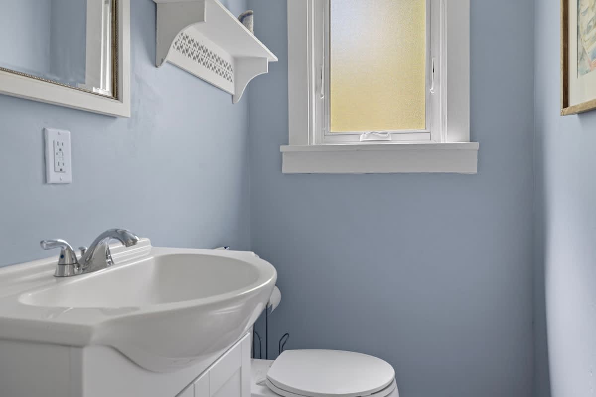 A compact bathroom with light blue walls, a white pedestal sink with a chrome faucet, a toilet, and a small window with frosted glass. A white shelf is mounted above the toilet, and a mirror hangs above the sink.