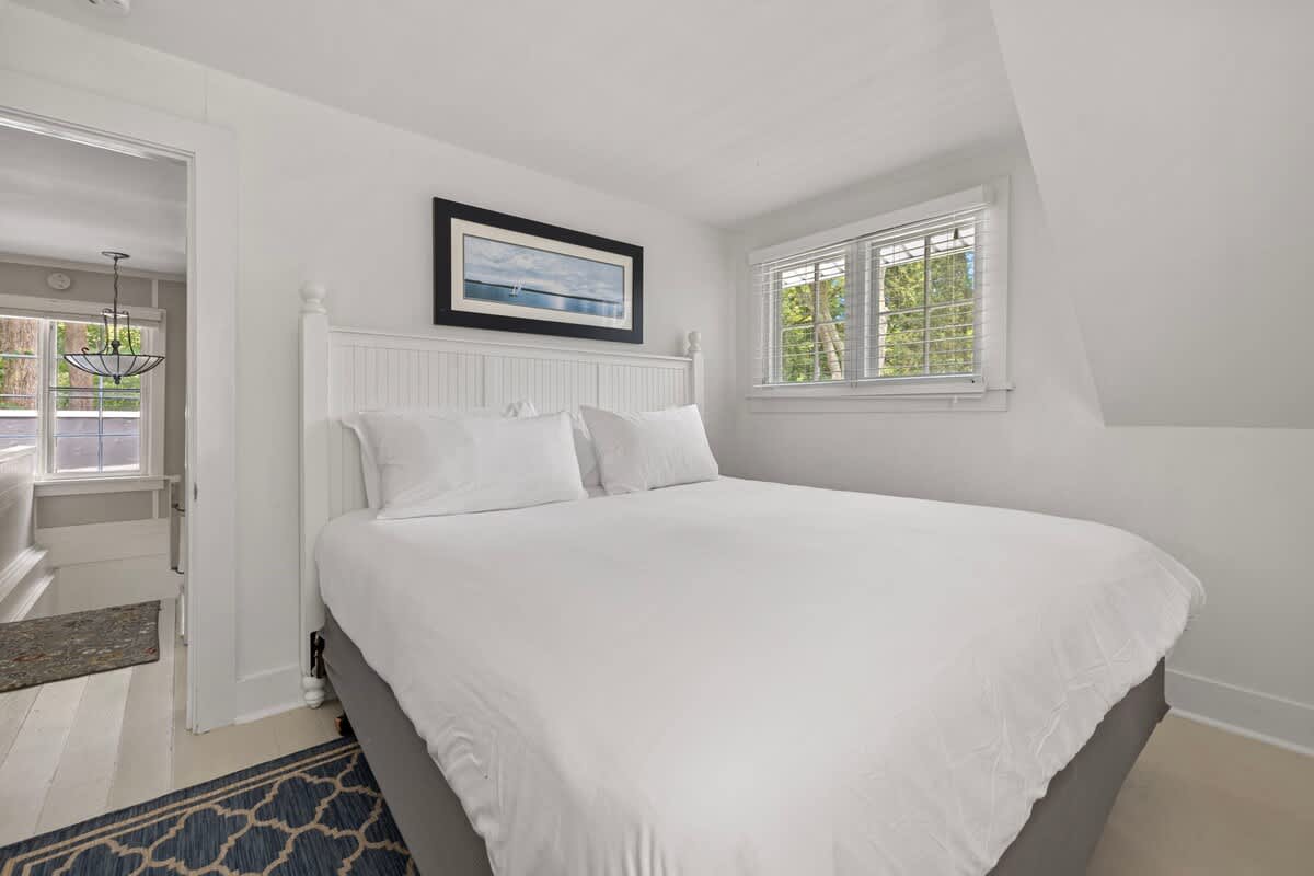 A bright bedroom with white walls and a white queen-sized bed featuring a slatted headboard and white bedding. A large window with white blinds overlooks lush greenery, and a framed seascape artwork hangs above the bed.