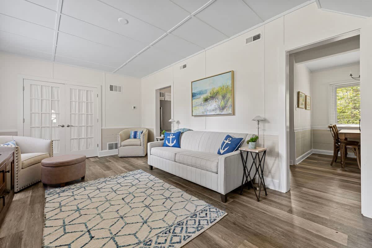 A bright living room features light wood-look flooring, white paneled walls, and a light gray sofa adorned with blue anchor pillows. A patterned rug anchors the seating area, with two armchairs and a wooden side table completing the space.