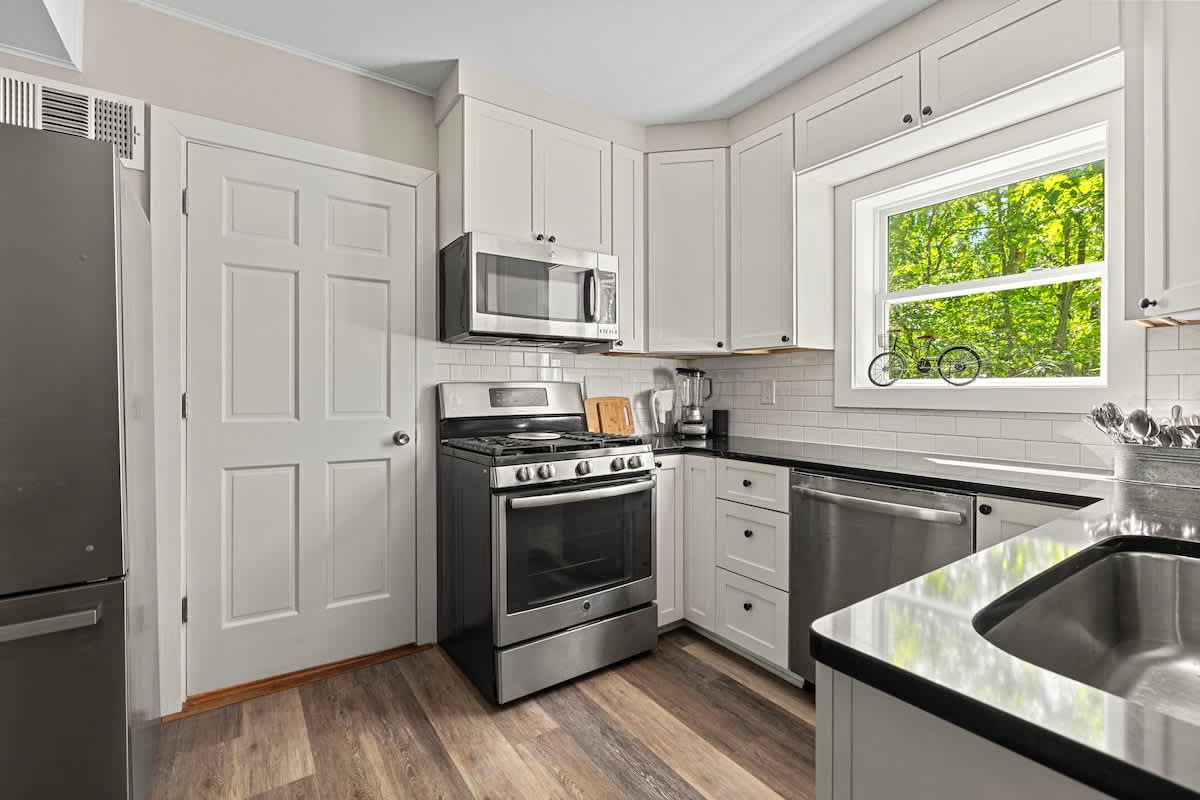 The bright kitchen showcases white shaker cabinets, dark countertops, and a white subway tile backsplash, with a stainless steel gas range and microwave. A closed white door is visible on the left, and a window above the sink offers natural light.