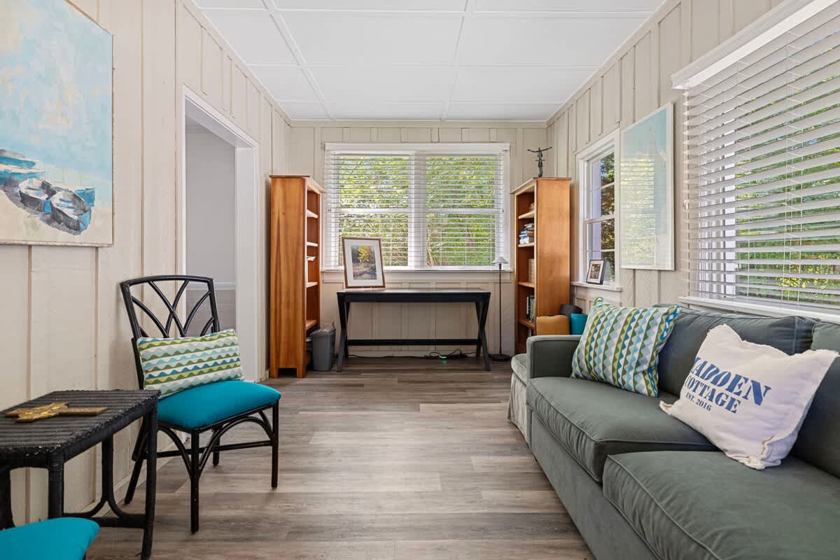 Cozy living room featuring a gray sofa with patterned pillows, light wood plank flooring, and a large window with blinds. Two wooden bookshelves flank a desk, and a black chair with a blue cushion is visible.