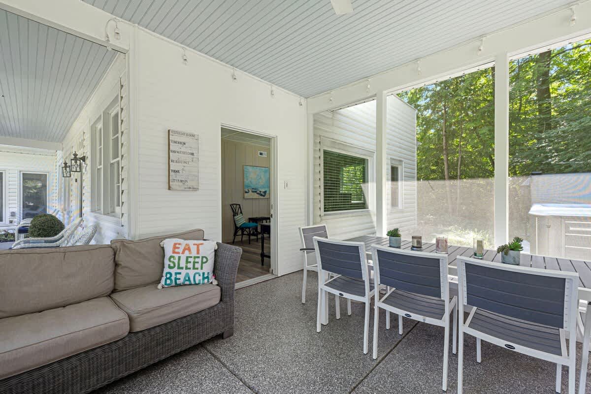 A screened-in porch with a dining area featuring a white table and grey chairs, and a wicker sofa with light cushions. The space offers views of the surrounding trees and includes a decorative 'EAT SLEEP BEACH' pillow.