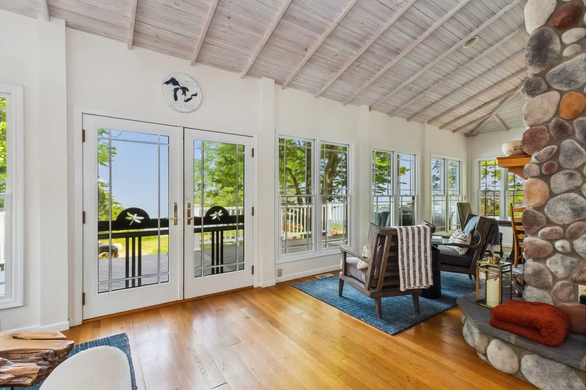 Cozy living area with light hardwood floors and a white vaulted ceiling with exposed beams. Large windows and glass French doors offer bright views of lush greenery and the lake, with comfortable armchairs arranged around a stone fireplace.