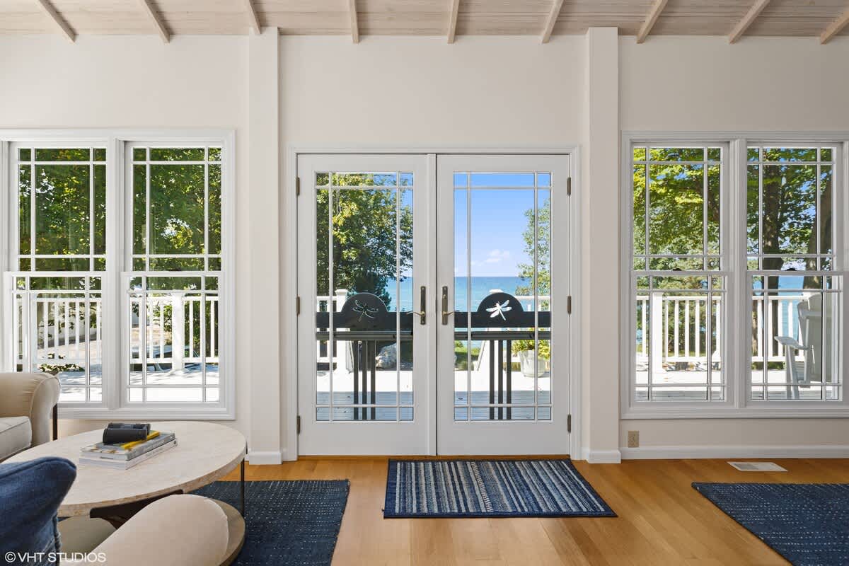 A bright living space features large windows and French doors with decorative dragonfly accents, offering expansive views of the lake and outdoor deck. The room has light wood floors, white walls, and a blue striped rug.