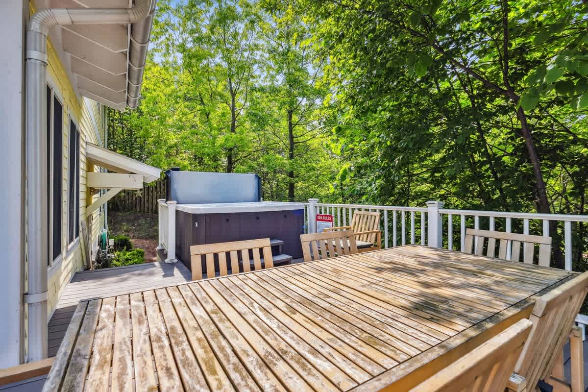 Wooden outdoor deck featuring a large hot tub and a rustic wooden dining table with chairs, surrounded by lush green trees. The deck is enclosed by white railings, providing a private space for relaxation and dining.