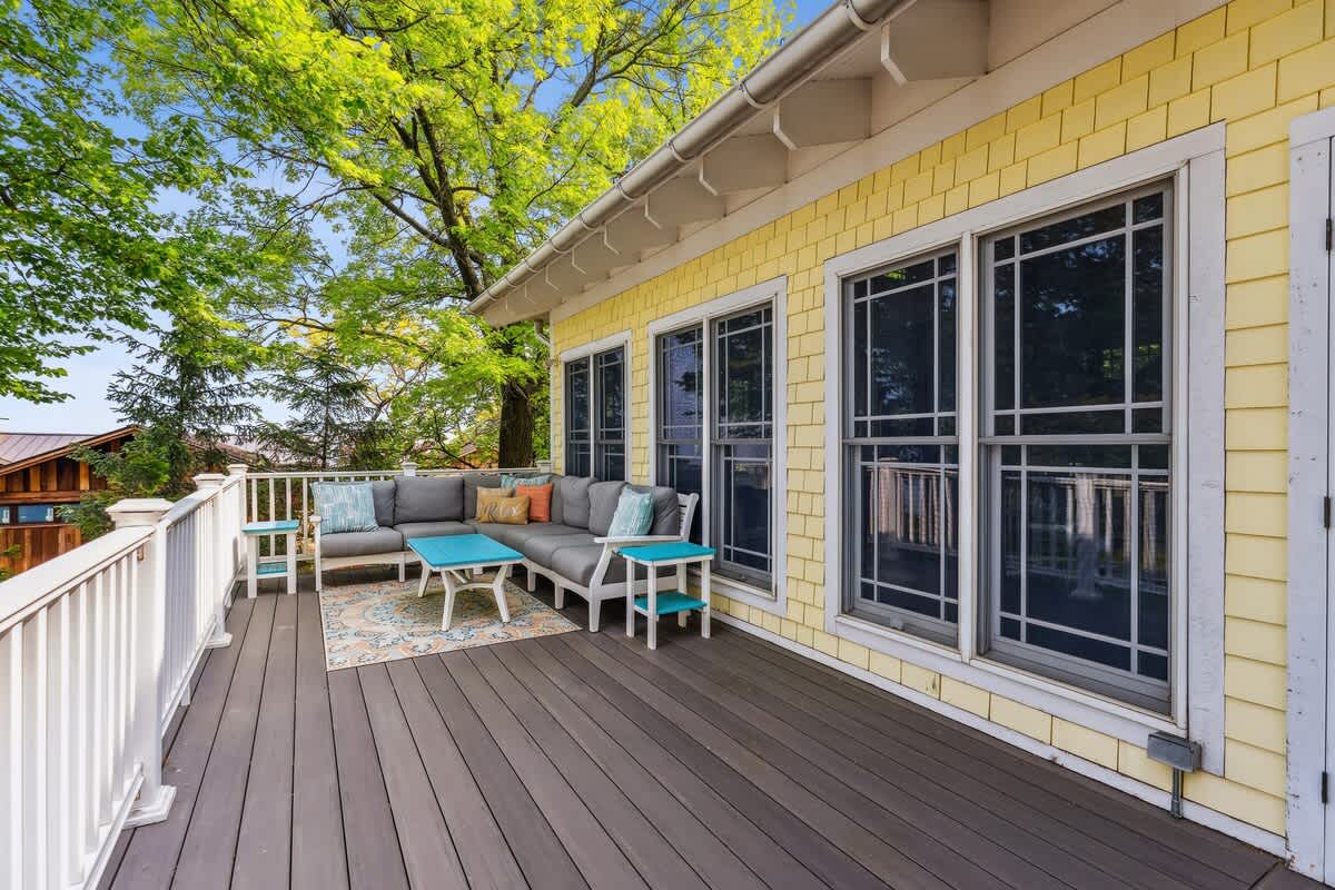 Expansive outdoor deck with a grey sectional sofa and blue accent tables, set on a patterned outdoor rug. The house features yellow shingle siding and large windows, surrounded by lush green trees.