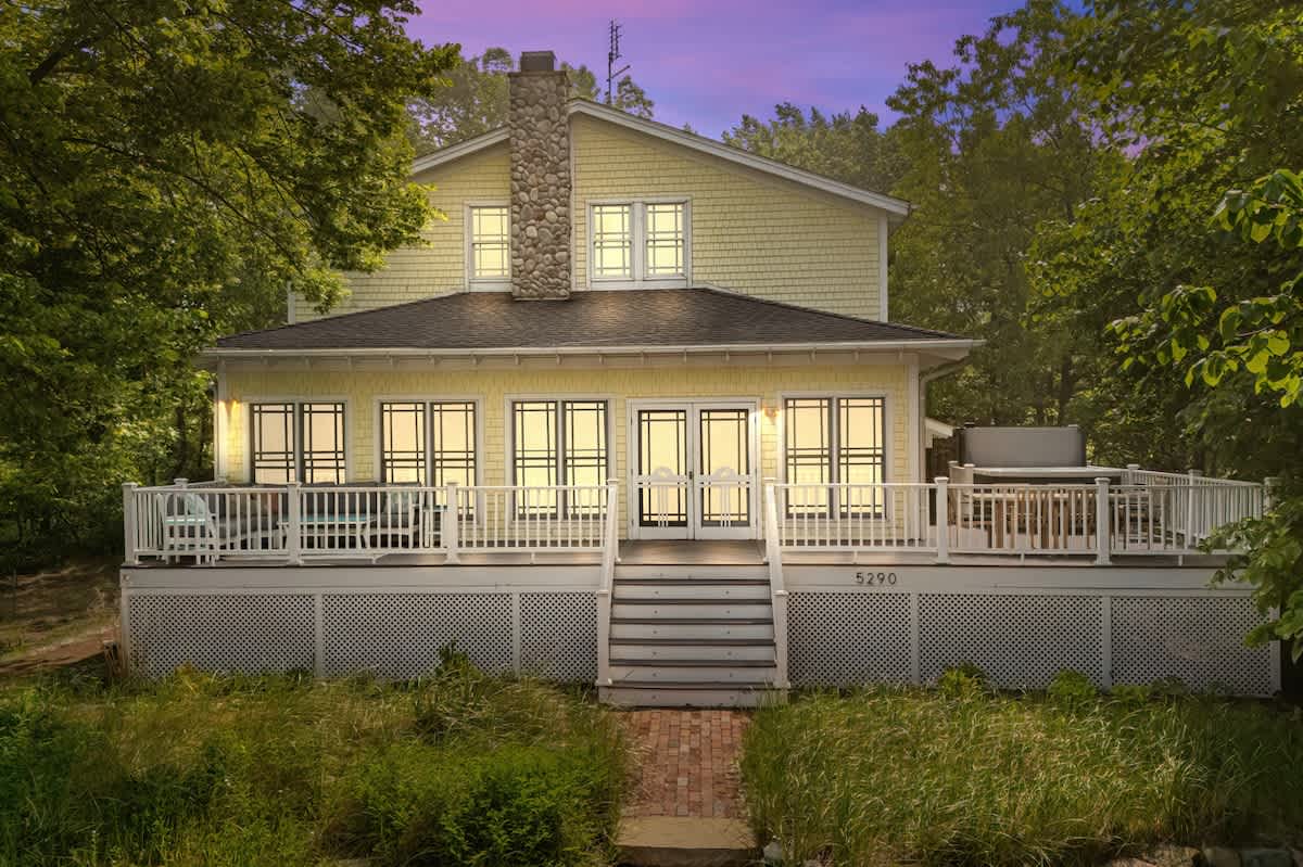 Rear exterior of a charming yellow house with white trim and a prominent stone chimney, featuring a large deck with white railings. Steps lead down from the deck to a grassy area, with lush trees providing a natural backdrop.