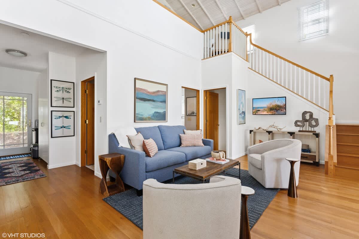 Bright living room with a blue sofa, two armchairs, a wooden coffee table on a blue rug, and a staircase leading to a loft area. Hardwood floors and white walls create an airy feel.