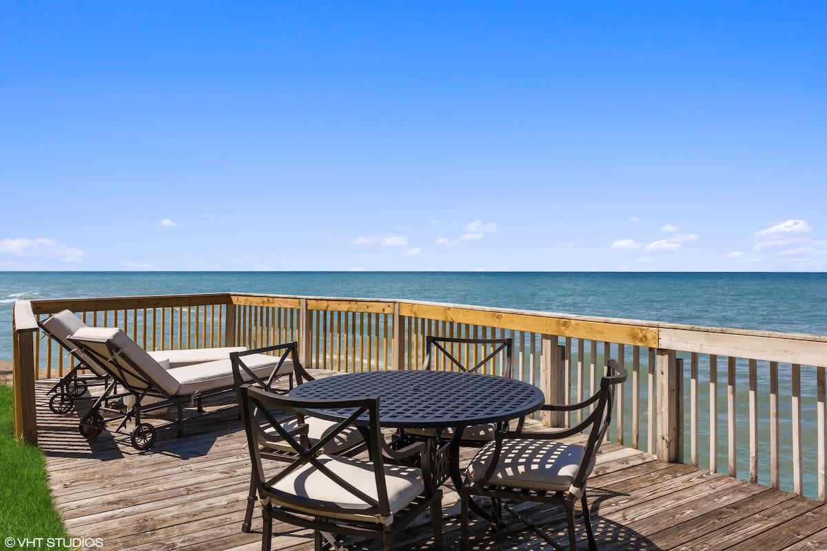 A spacious wooden deck overlooks a serene blue lake under a clear sky, featuring a black metal outdoor dining set with four chairs and two lounge chairs. A white railing encloses the deck, providing unobstructed water views.