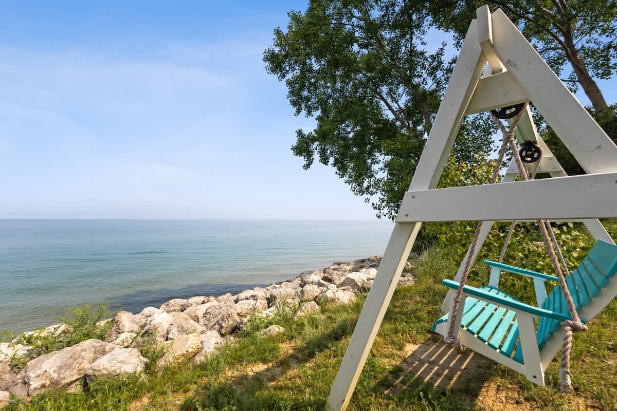 A-frame swing set with a blue wooden swing, positioned on a grassy slope overlooking a serene lake under a clear blue sky. Lush green trees frame the tranquil water view.