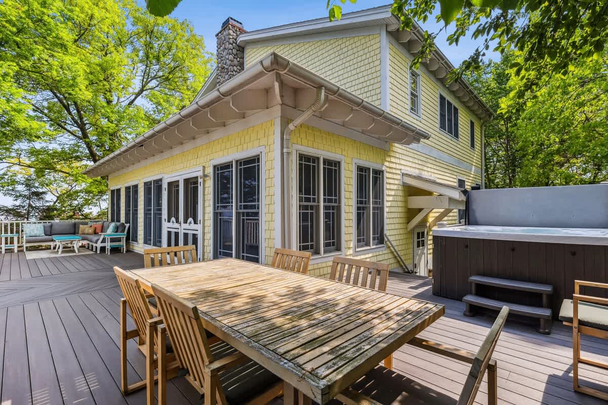 A spacious outdoor deck features a large wooden dining table with chairs, a hot tub, and a comfortable outdoor sofa seating area. The yellow house exterior is visible, surrounded by vibrant green trees under a sunny sky.
