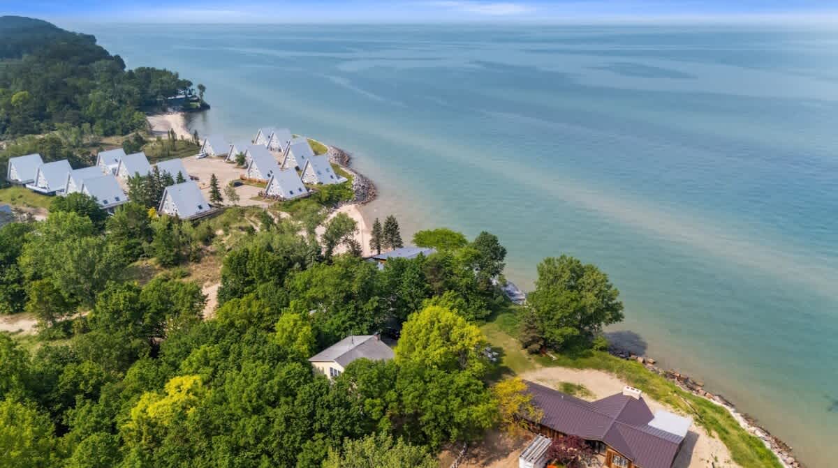 An expansive aerial view of a serene coastline with a wide blue lake, featuring several A-frame style houses and other properties nestled among abundant green trees along the water's edge, highlighting the community layout.