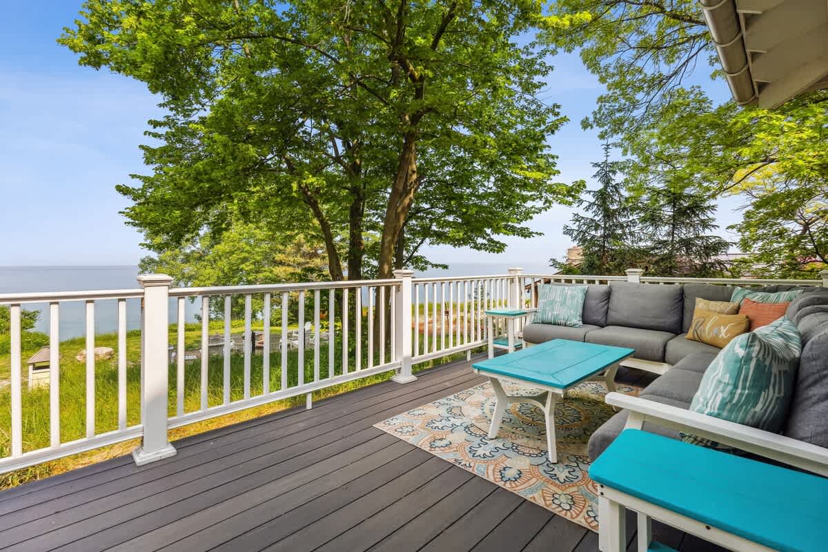 Spacious outdoor deck with dark composite flooring and white railings, furnished with a grey sectional sofa and a turquoise coffee table. The deck offers a beautiful view of the lake through surrounding green trees.