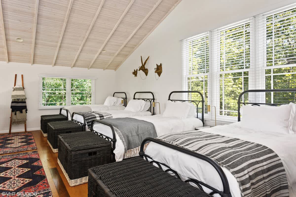 Spacious loft bedroom with four single beds featuring black metal frames and white bedding, each with a woven storage trunk at the foot. Large windows provide natural light, and the room has vaulted white ceilings.