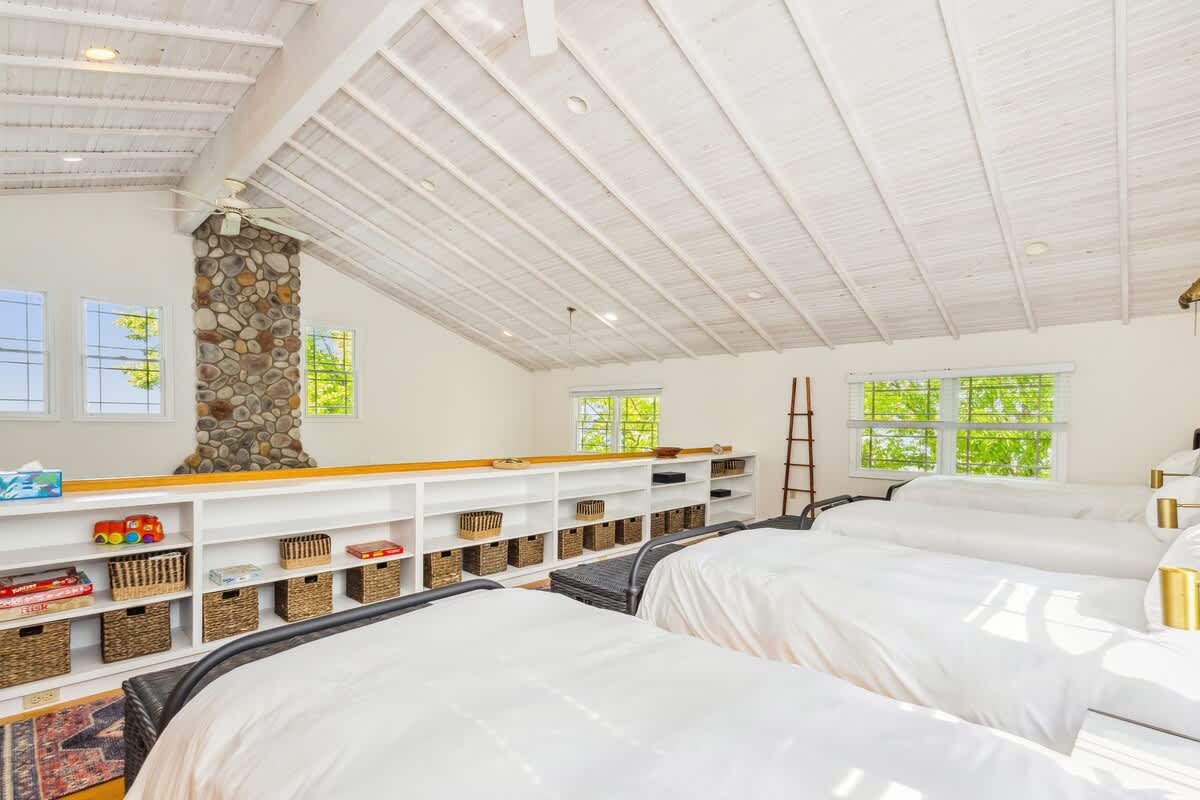 Wide view of the loft bedroom, showcasing four single beds, extensive built-in shelving with baskets, and the upper portion of the stone fireplace. The room features vaulted white ceilings and hardwood floors.