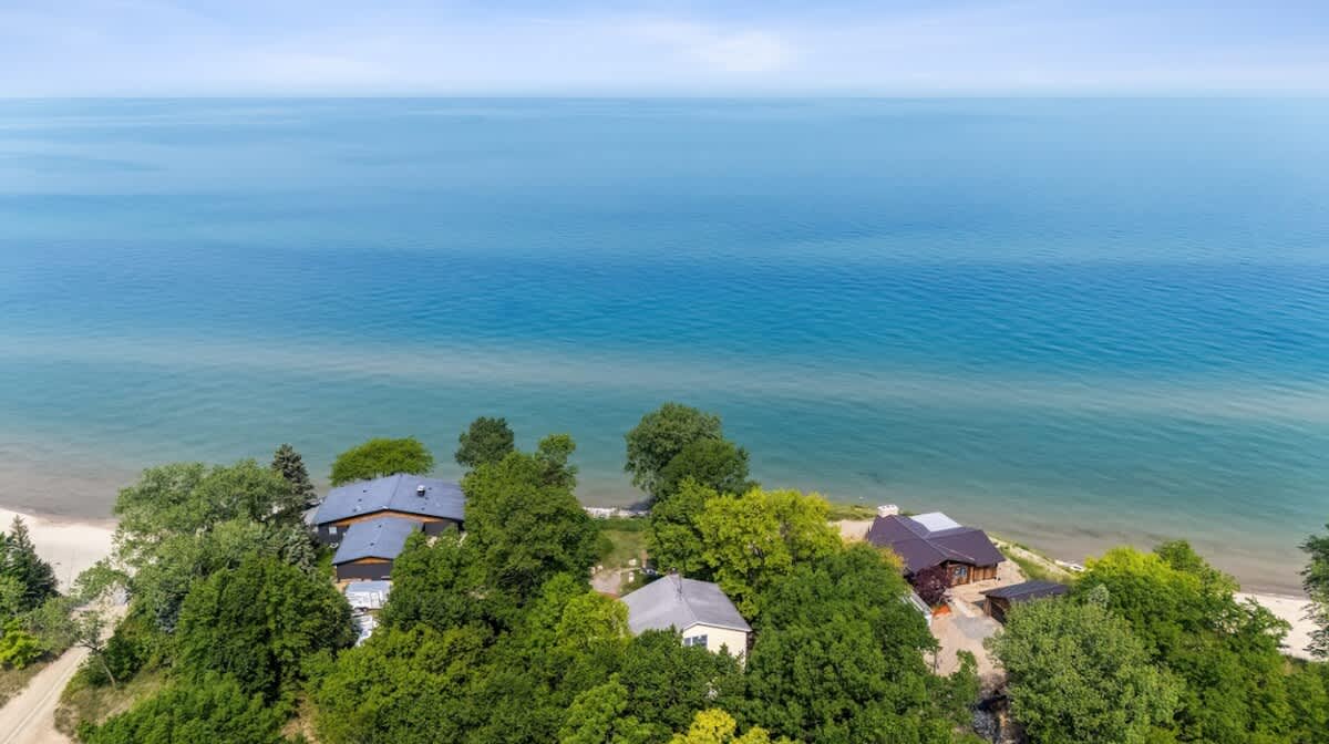 A high aerial view showcasing the vast expanse of a calm, blue lake or ocean meeting the shoreline, with a few houses and dense green trees along the coast under a clear sky, emphasizing the property's lakeside location.