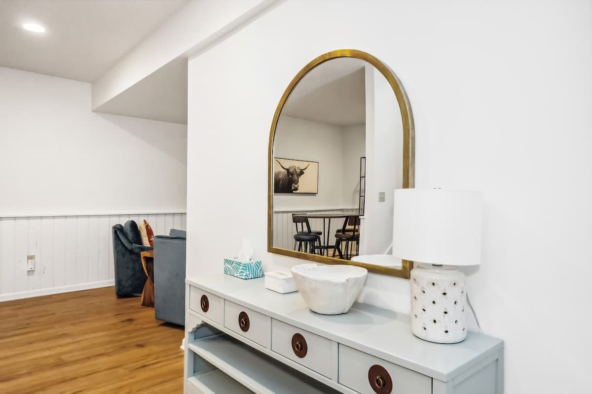 Bright entryway featuring a light grey console table with drawers, topped by a large arched gold-framed mirror. A white lamp and decorative bowl sit on the table, with a glimpse of the living area beyond.