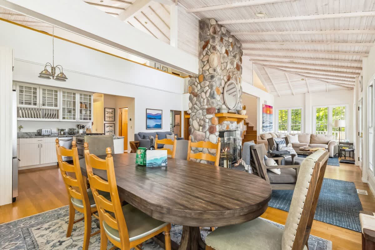 Open-concept living space showcasing a kitchen with white cabinets, a dining area with a dark wood table, and a living room with a sofa, all centered around a large stone fireplace. Hardwood floors connect the areas.
