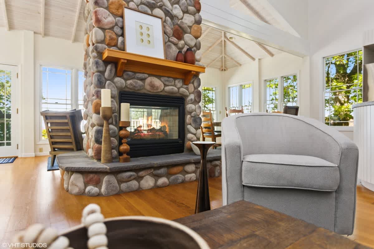 A cozy living room features a prominent floor-to-ceiling stone fireplace with a wooden mantel, a light gray armchair, and warm hardwood floors. Large windows allow natural light and offer glimpses of the surrounding greenery.