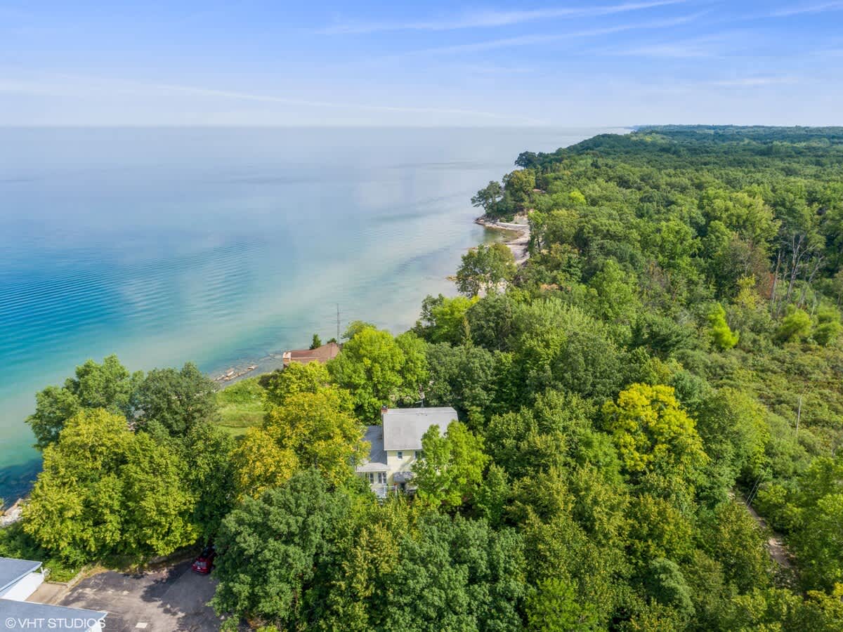 An aerial view showcases a yellow lakeside house nestled among dense green trees, with the vast blue expanse of the lake stretching to the horizon. The property is surrounded by lush forest, highlighting its secluded, natural setting.