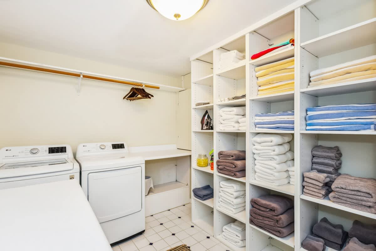 Bright laundry room with white tiled floors and ample shelving for linens and towels. It features a white washer and dryer, a hanging rod, and an ironing board, all under a flush-mount ceiling light.