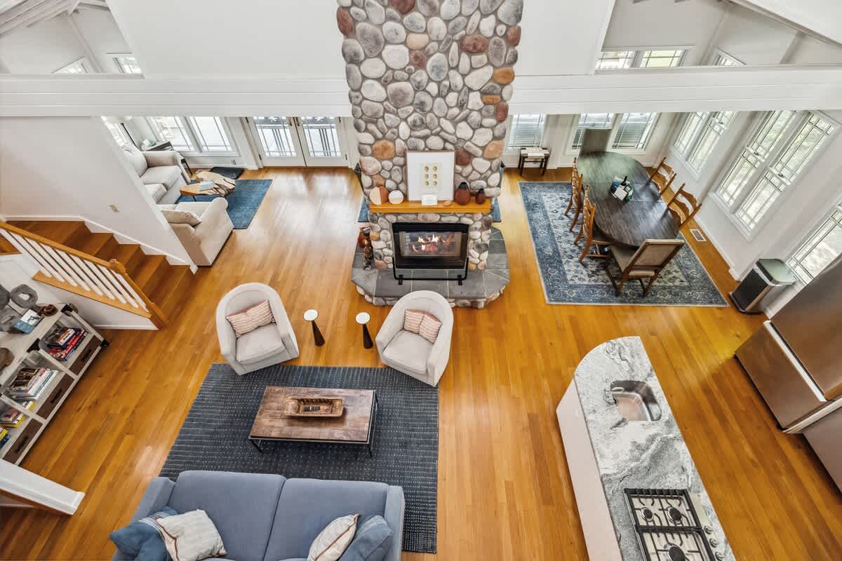 Expansive open-concept living space with light hardwood floors, a towering stone fireplace, and a high white ceiling. The area includes a living room with blue rugs, a dining area with a large wooden table, and a partial view of the kitchen island.