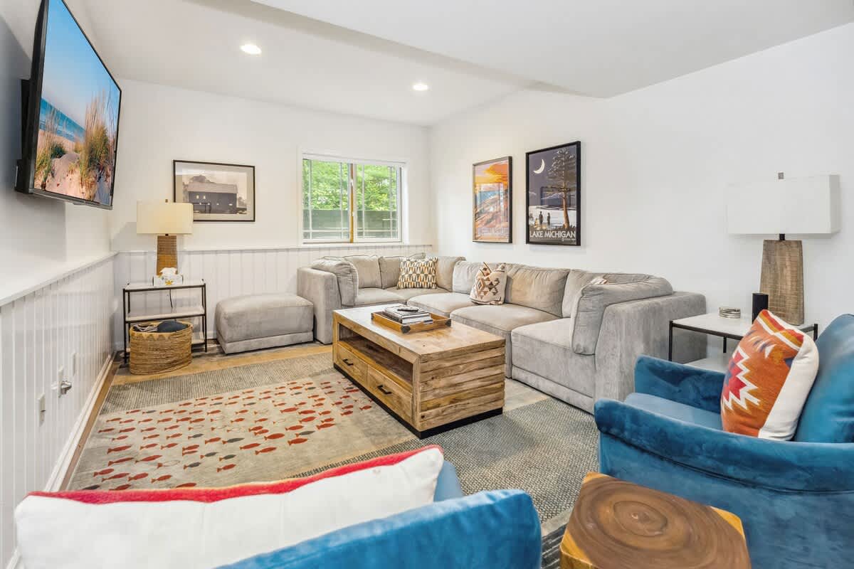 Spacious living room with a large grey sectional sofa, a rustic wooden coffee table, and a wall-mounted TV. White wainscoting lines the lower walls, and a patterned rug covers the floor, complemented by blue accent chairs.