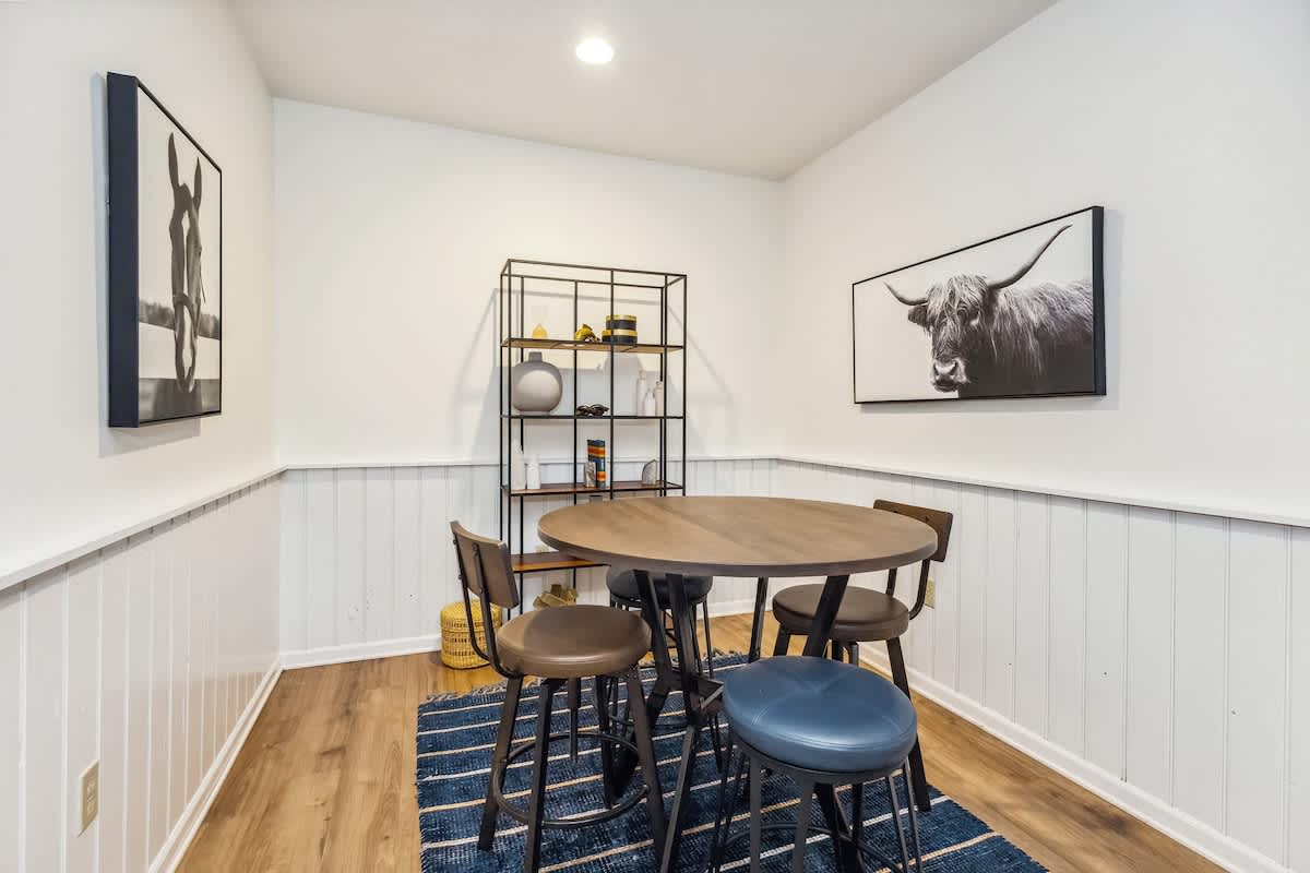Inviting dining area featuring a round wooden table with four stools, set on a blue striped rug. White wainscoting lines the walls, which are adorned with framed artwork, and a black metal shelving unit holds decorative items.