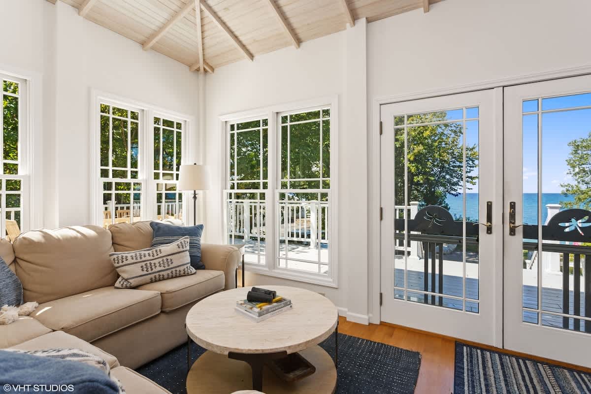 A bright living room features a beige sofa with blue throw pillows, a round marble coffee table, and large windows and French doors offering stunning lake views. The room has light wood floors and a vaulted ceiling with exposed white beams.