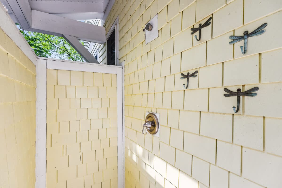 An outdoor shower area featuring yellow shingle siding, equipped with a modern bronze shower fixture and decorative dragonfly-shaped hooks for towels, partially enclosed with white trim.