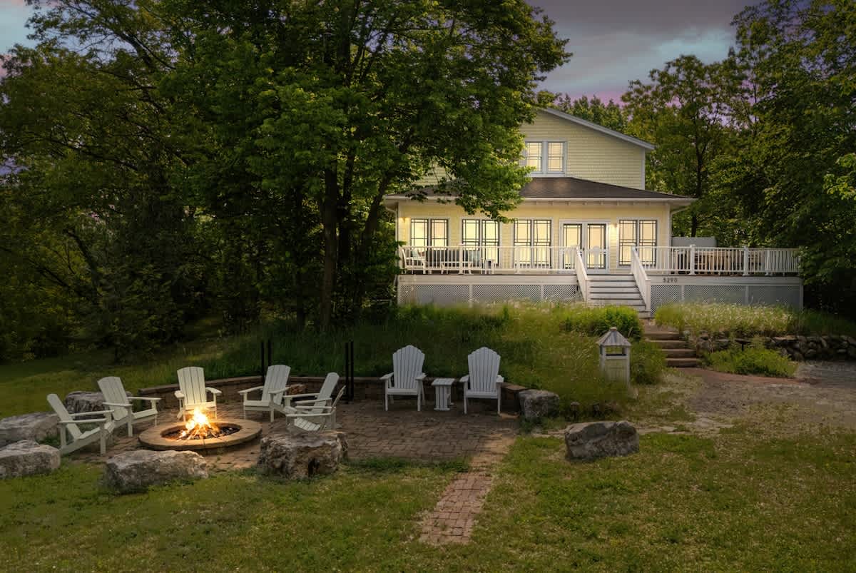 The back of a yellow house with white trim and a large deck is illuminated at dusk, leading down to a brick patio with a lit fire pit and white Adirondack chairs. Lush green trees and tall grasses surround the property, with the lake visible in the background.