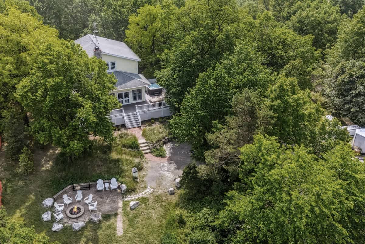 An aerial view of a yellow house nestled among dense green trees, featuring a multi-level deck with a hot tub and a stone fire pit area with white Adirondack chairs in the foreground, all surrounded by lush foliage.