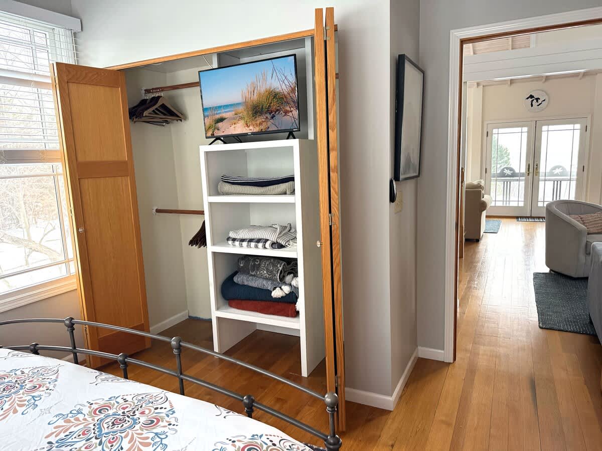 Open closet area within a bedroom, featuring bi-fold wooden doors, shelving with folded linens, and a television mounted above. Hardwood floors and light gray walls are visible.