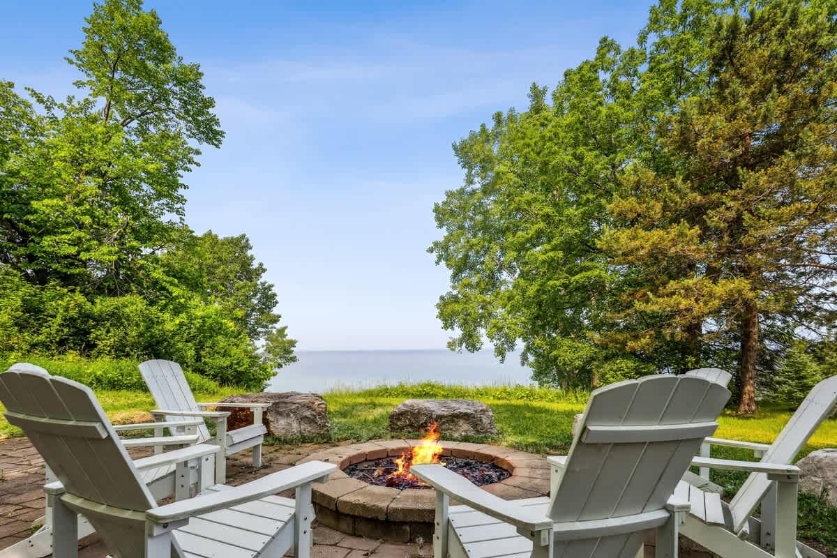 Outdoor fire pit area with a stone-lined fire pit, surrounded by four white Adirondack chairs on a paved patio. Lush green trees frame a distant view of a calm lake under a clear blue sky.