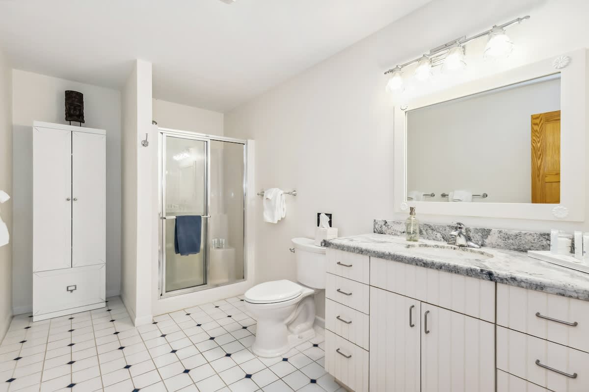 Bright bathroom featuring white tiled floors with black accents, a glass-enclosed shower, toilet, and a white vanity with a grey granite countertop and mirror. A tall white storage cabinet is visible on the left.