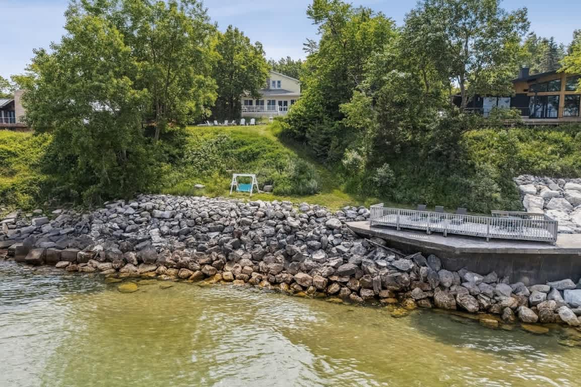 Aerial view of a yellow house nestled among green trees, overlooking a rocky shoreline and a calm lake. A wooden deck extends over the water, and a swing set is visible on the grassy slope leading to the shore.