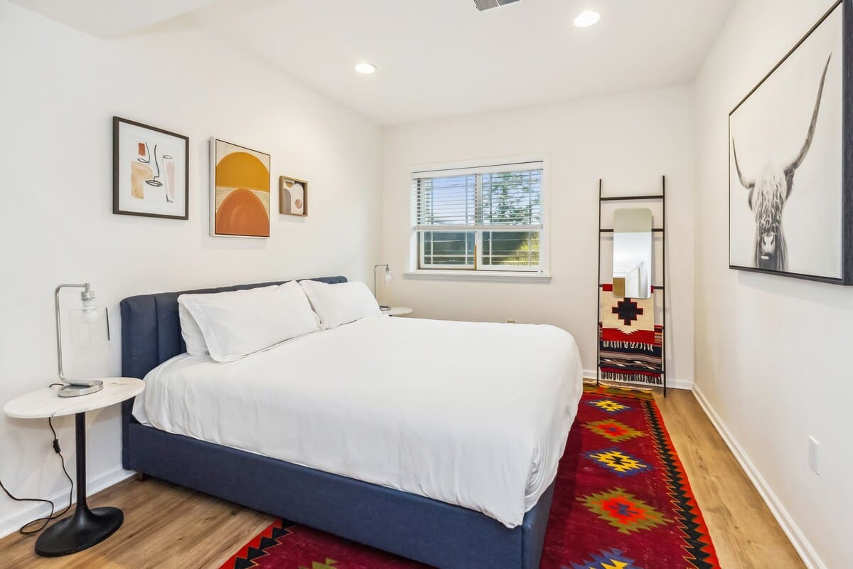 Modern bedroom with white walls, a blue upholstered queen bed, and a window with white blinds. A vibrant red and black patterned rug covers the light wood floor, and abstract art adorns the walls.