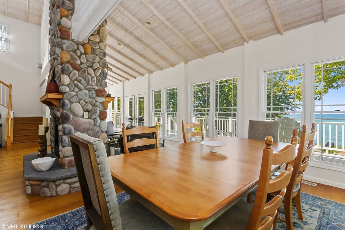 Spacious dining area featuring a large wooden table with seating for eight, set on a blue patterned rug. A prominent stone fireplace anchors the room, with large windows offering lake views.