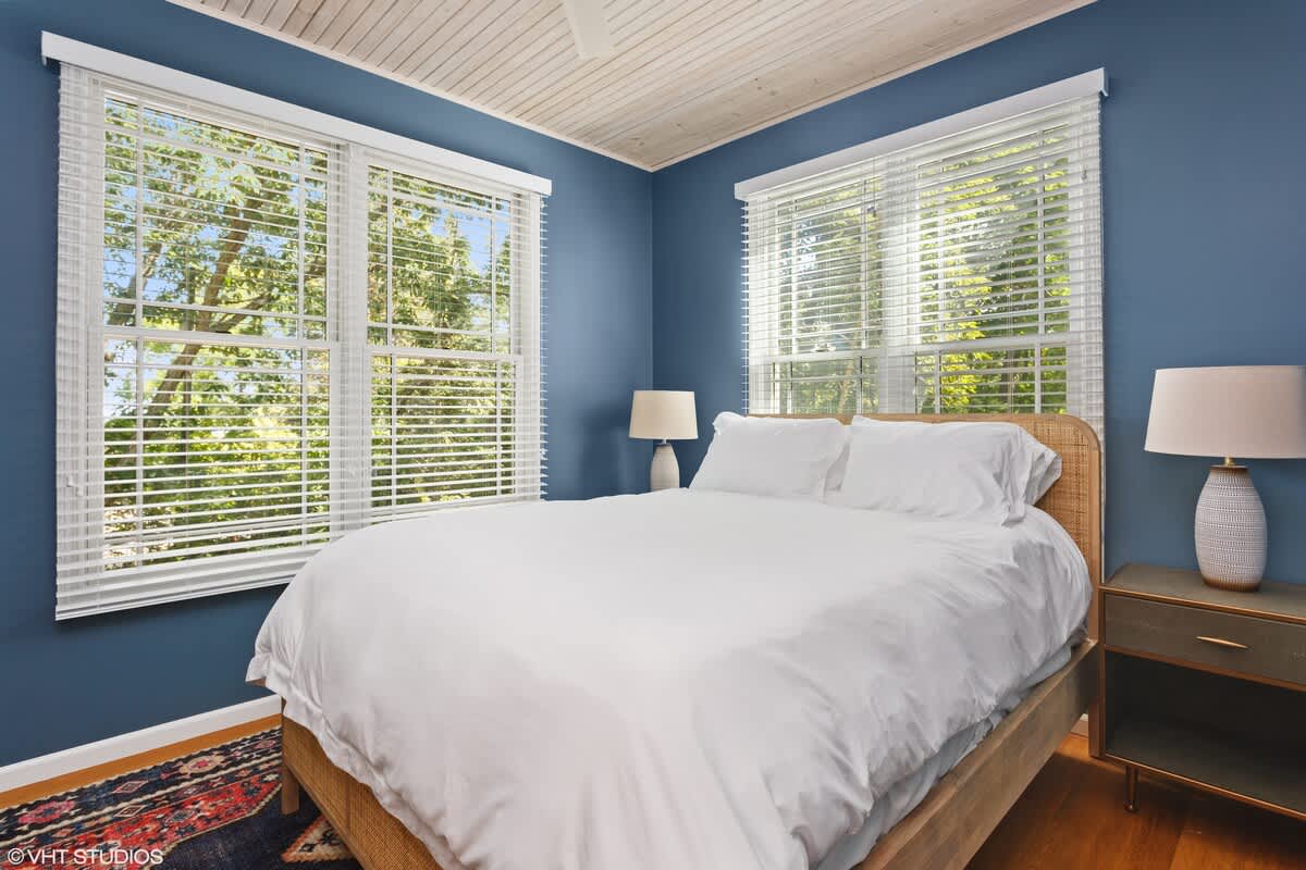 Cozy bedroom with vibrant blue walls, a white queen-sized bed with a woven headboard, and two large windows with white blinds offering views of lush greenery. The ceiling is paneled wood.