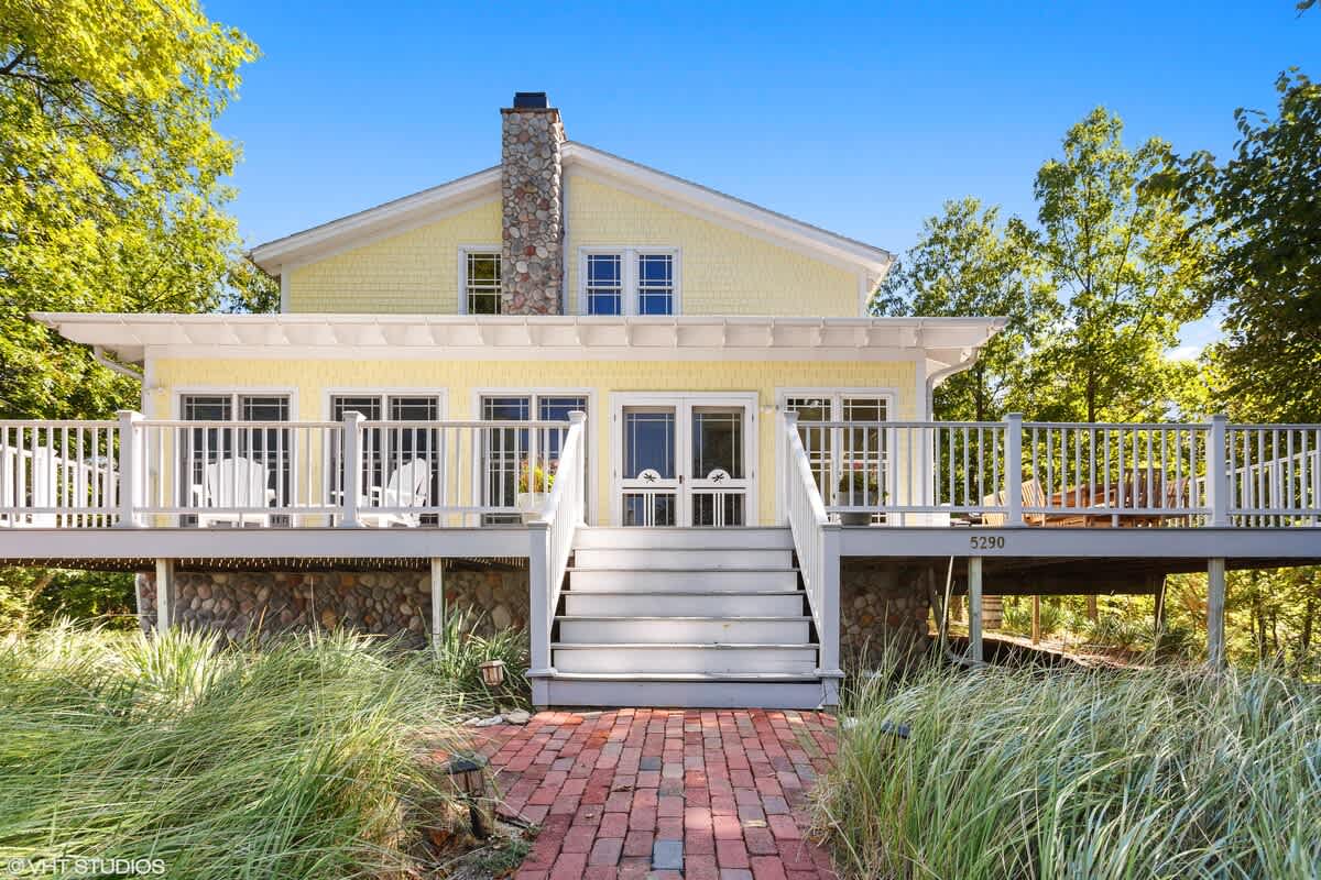 The front exterior of a charming yellow house with white trim features a large elevated deck accessible by a wide white staircase. A stone chimney is visible, and the property is surrounded by lush greenery and tall grasses.