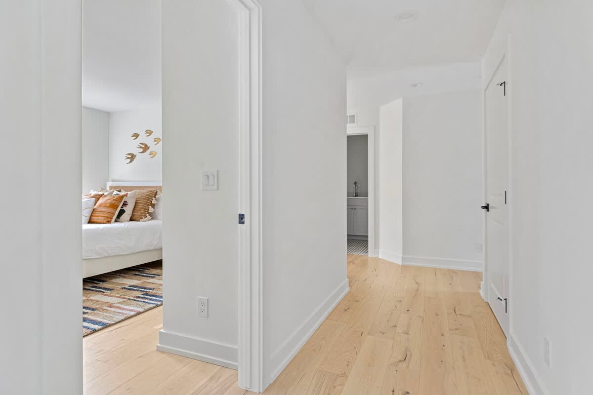 A bright hallway with light hardwood floors and white walls, providing a partial view into a bedroom with a patterned rug and decorative pillows, and another doorway further down the hall.