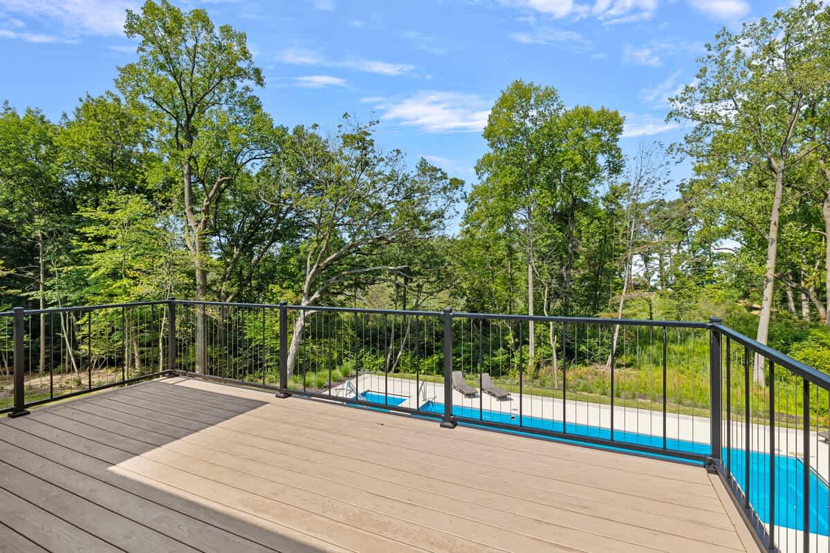 A spacious outdoor deck with light-colored composite decking and a black metal railing, offering an elevated view of a bright blue swimming pool surrounded by lush green trees under a clear sky.