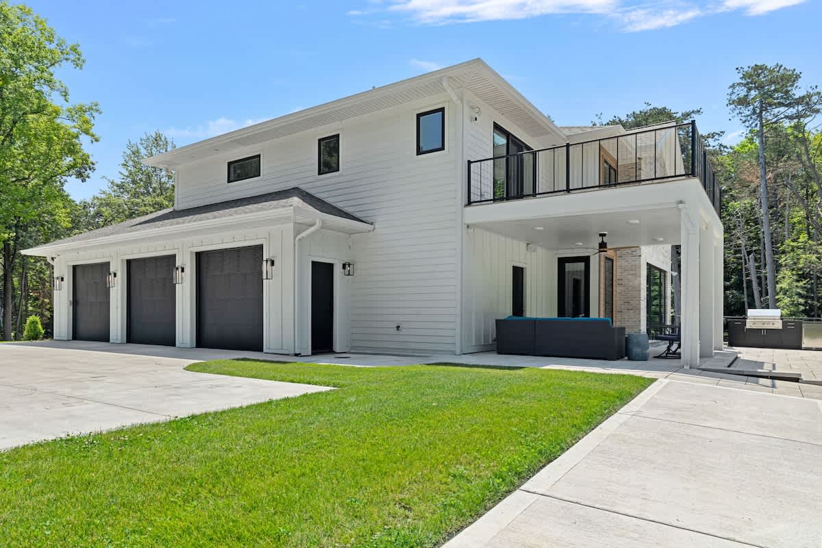 Side view of a contemporary white house with black-framed windows and a three-car garage, featuring a covered patio with outdoor seating and a second-story balcony with black railings. Lush green grass and trees surround the property.