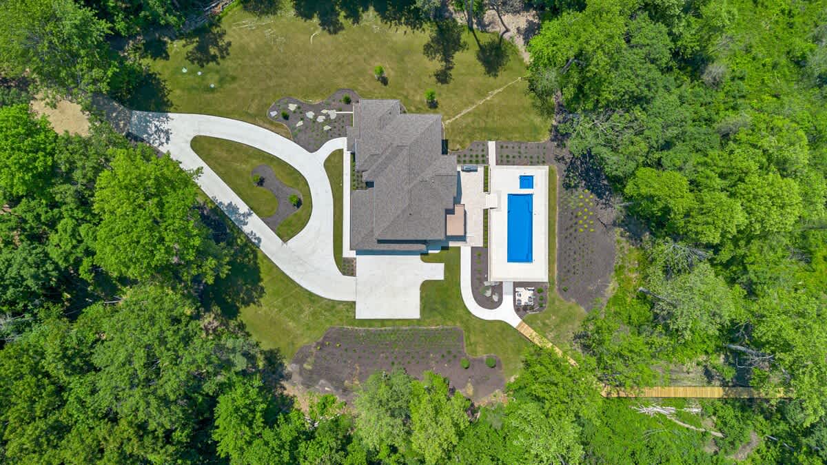 Overhead aerial view of a modern estate featuring a large house with a dark roof, a sweeping curved concrete driveway, a rectangular swimming pool, and extensive green landscaping, all nestled within a dense forest.