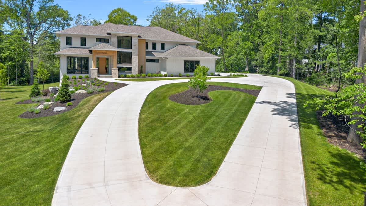 Front exterior view of a grand, modern two-story house with light-colored siding, dark-framed windows, and a prominent stone and wood entrance, set amidst a manicured lawn and a sweeping circular concrete driveway.