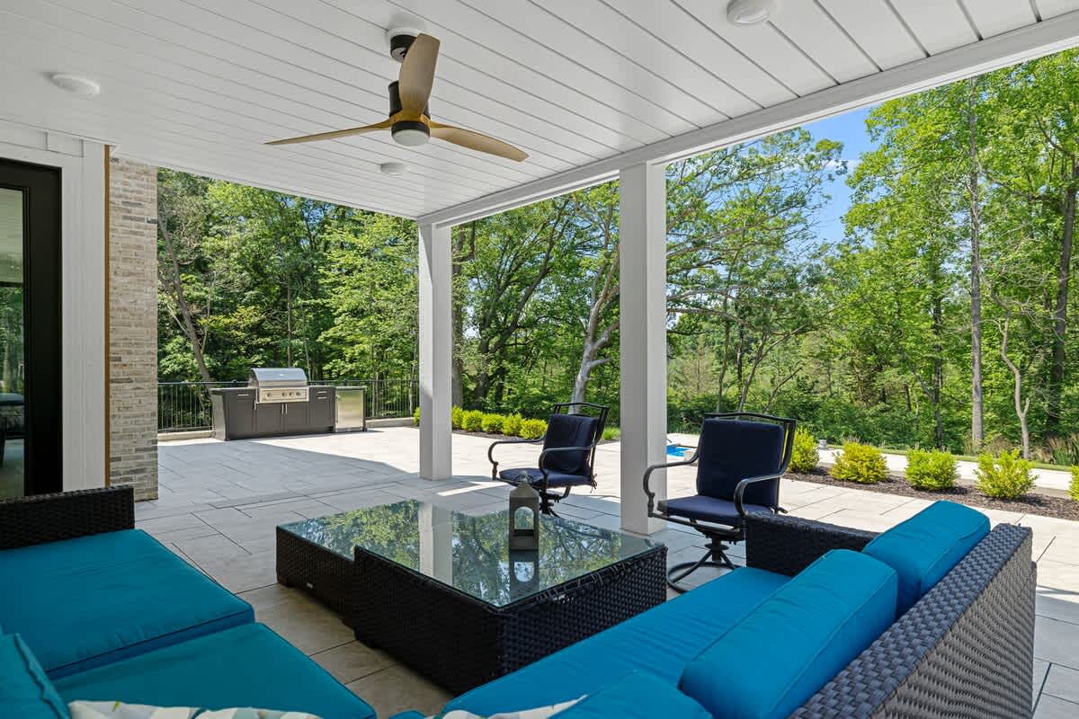 A covered outdoor patio area with comfortable wicker seating featuring bright blue cushions and a glass-top coffee table. A ceiling fan is mounted above, and the space overlooks a lush green landscape and a pool area.