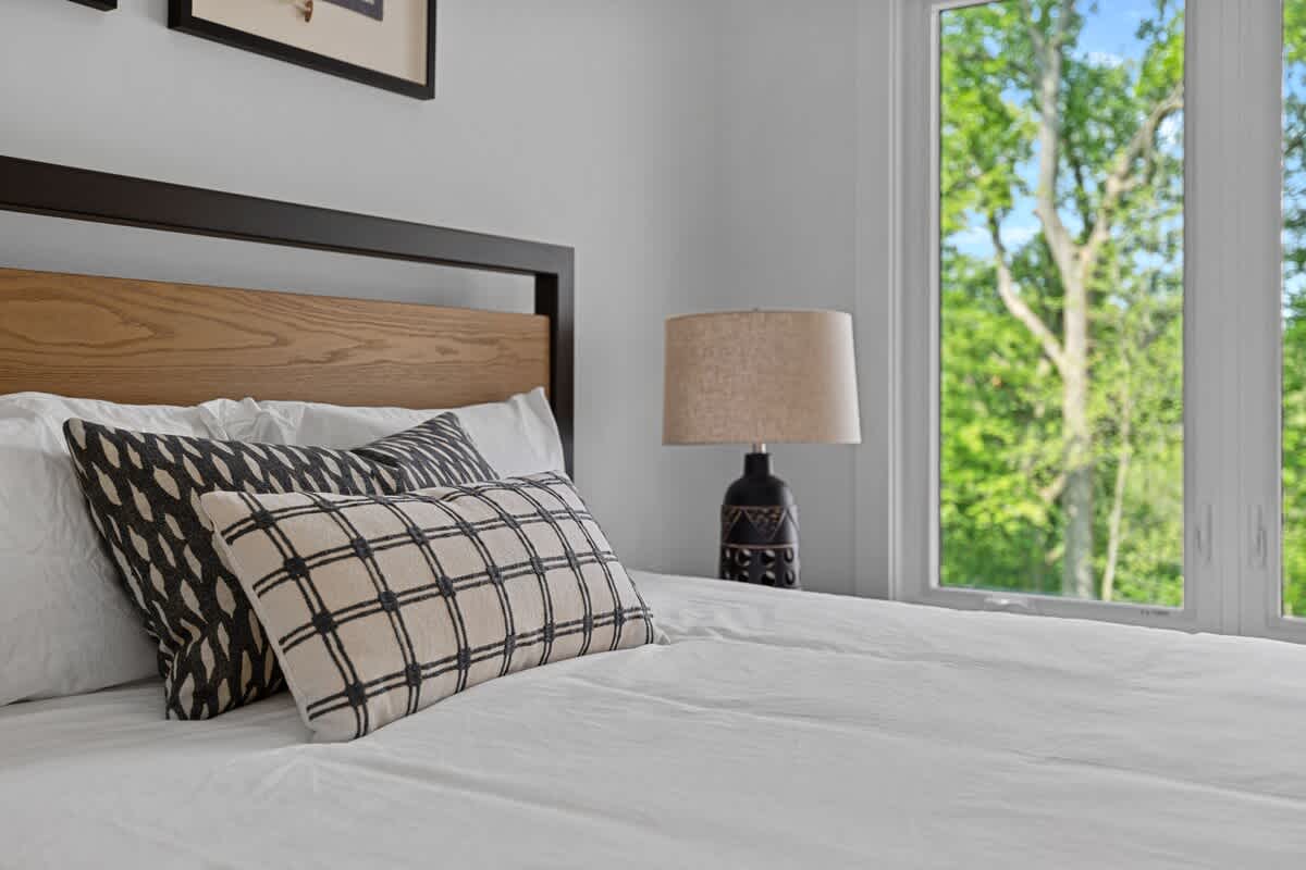 Close-up of a modern bed with white bedding and decorative pillows, including a black and white plaid one. A black nightstand with a textured black lamp and a beige shade sits beside the bed, next to a large window with a view of green trees.