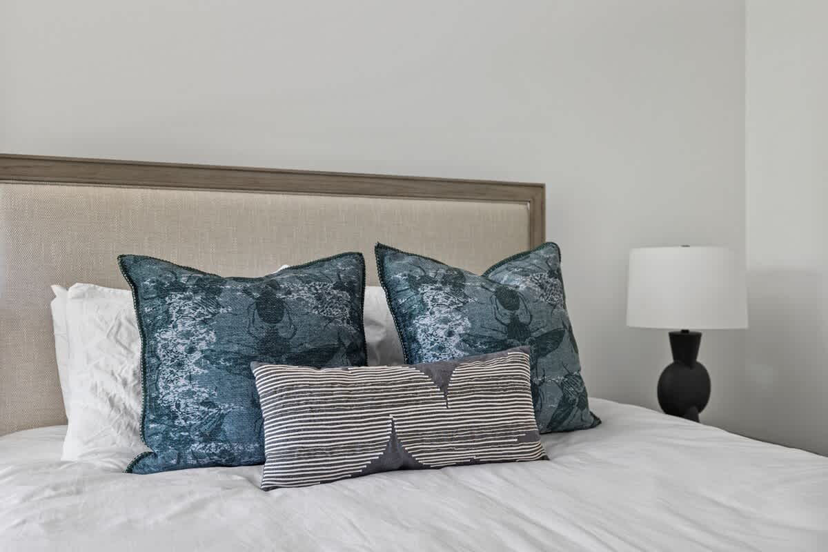 Close-up of a bed in bedroom-1 with white linens, two large teal patterned pillows, and a striped lumbar pillow, beside a modern black lamp on a nightstand.